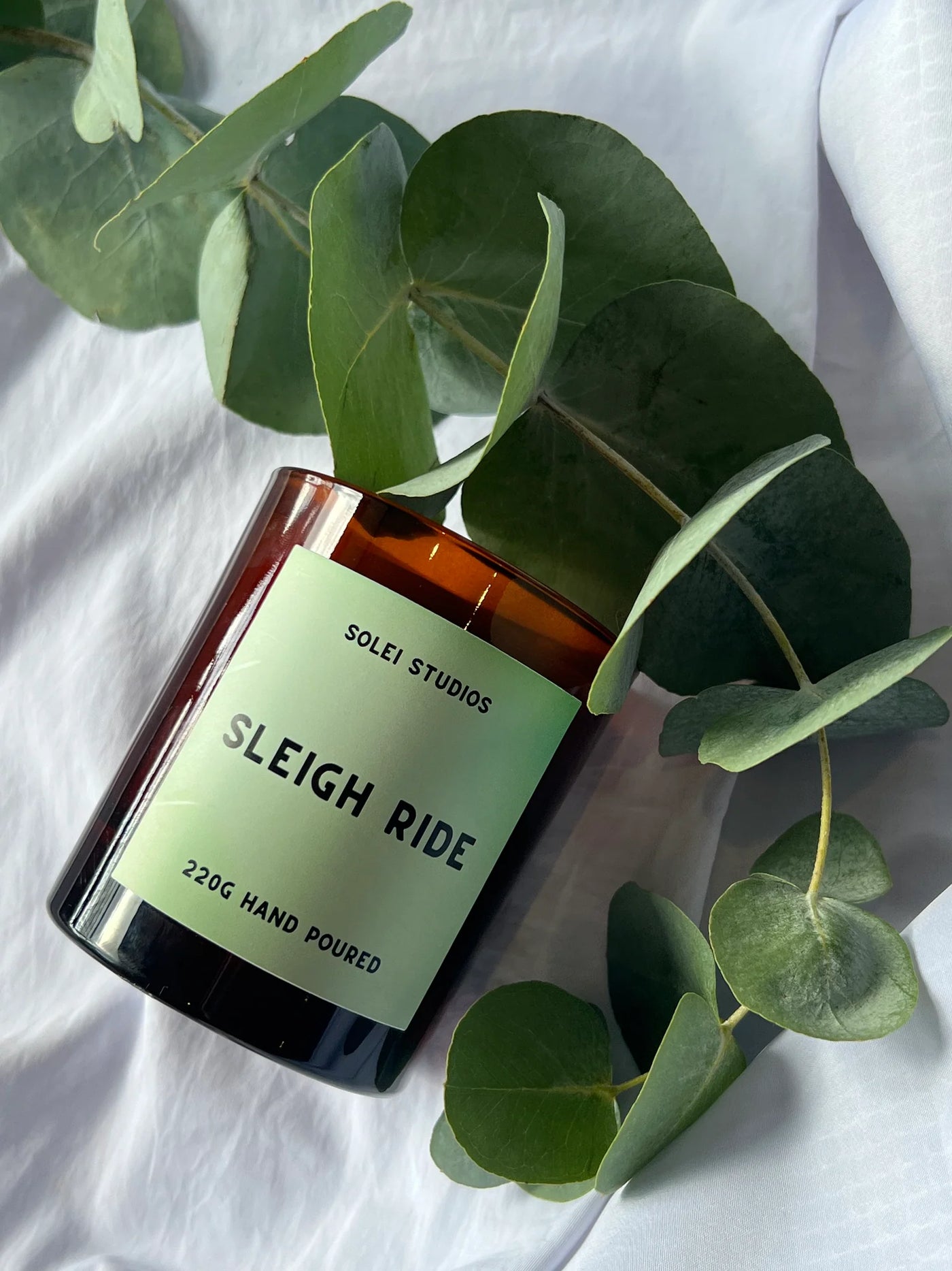 Scented candle labeled 'Sleigh Ride' from Solei Studios with eucalyptus leaves on a white background