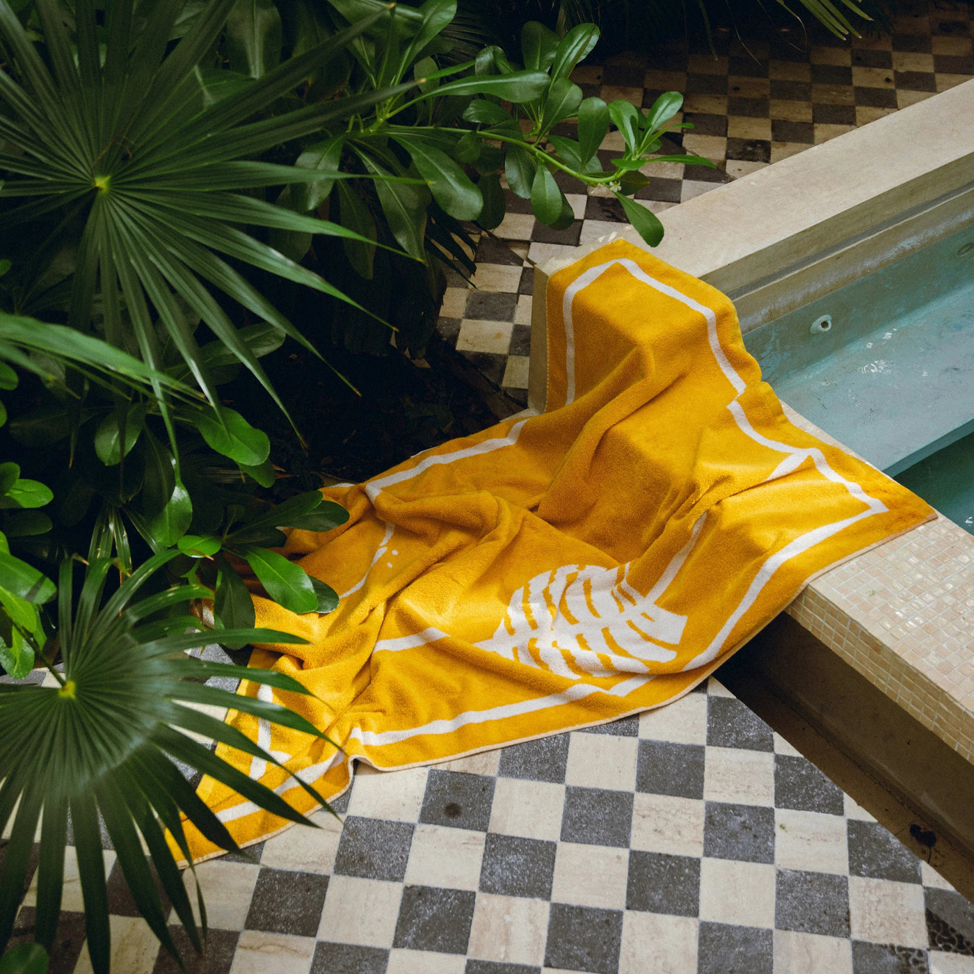 Yellow towel with a white design draped over a chair by a poolside with greenery.