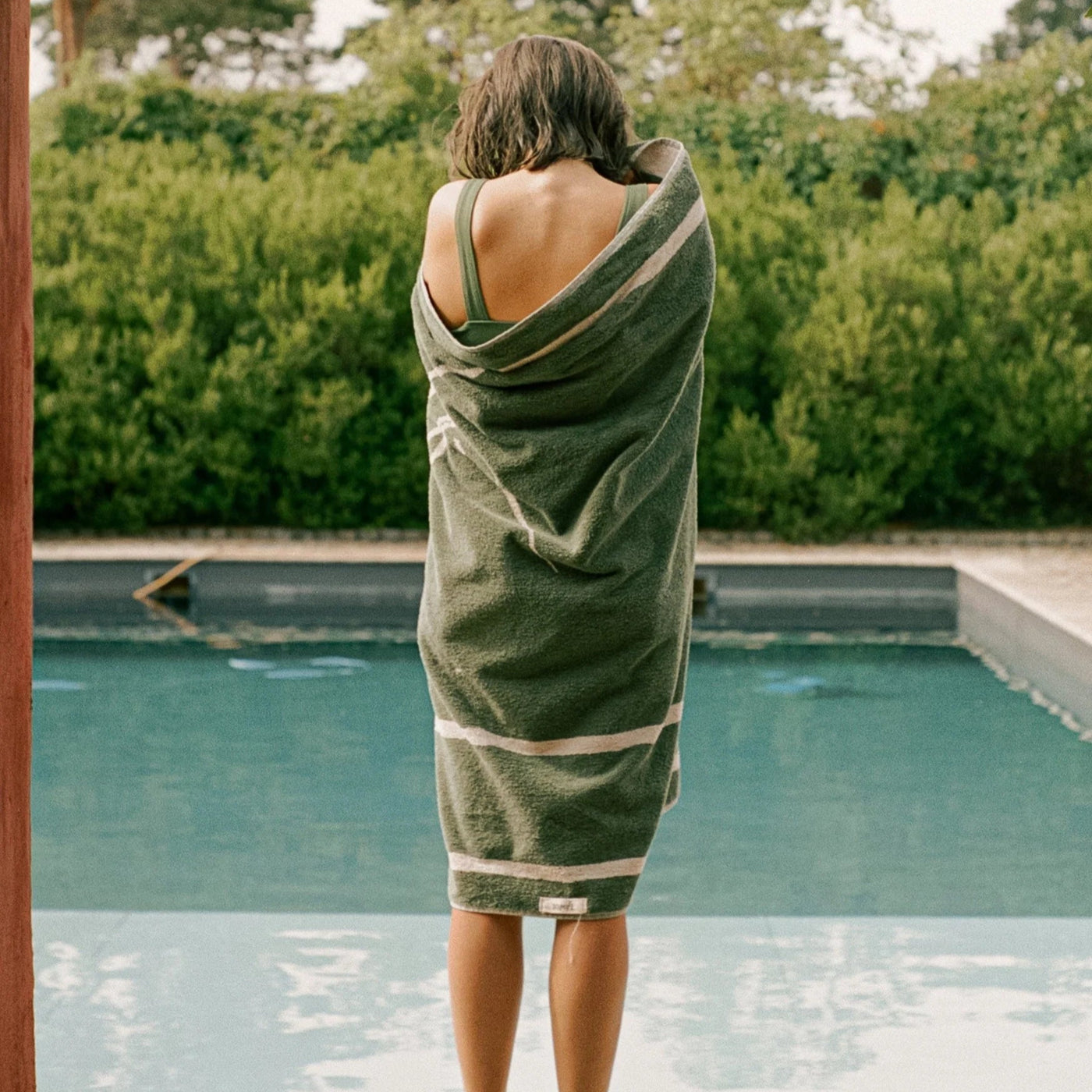 Person wrapped in a green towel by a pool with greenery in the background