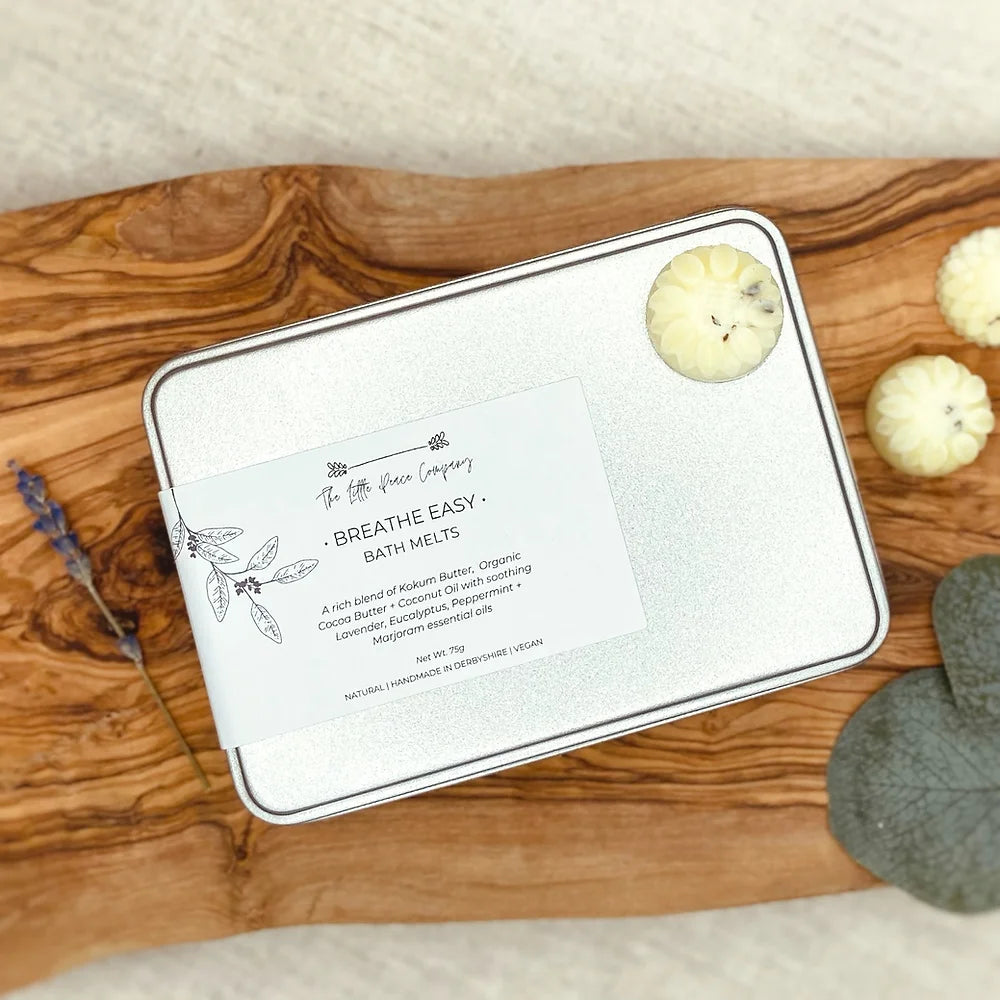 A tin of 12 bath melts by The Little Peace Company placed on a wooden surface, with individual melts visible alongside lavender and rosemary leaves.