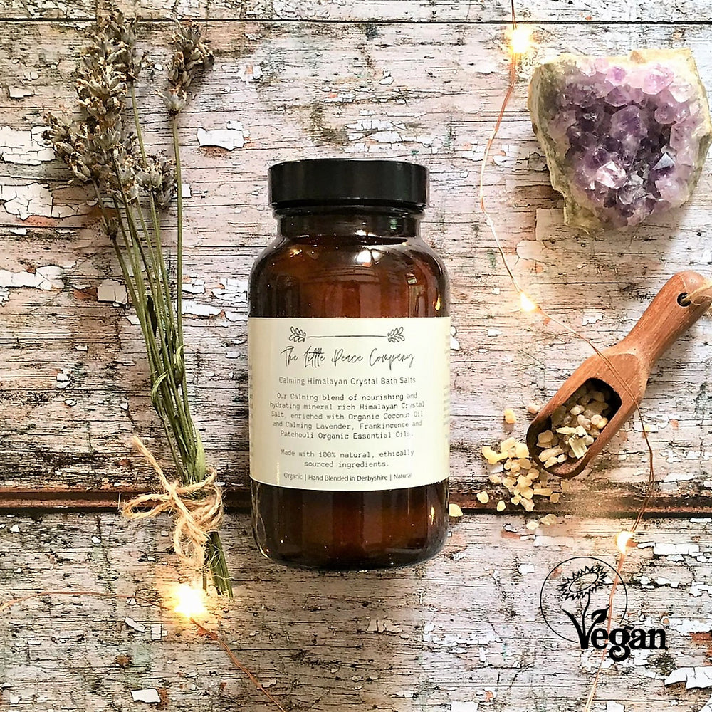 A jar of The Little Peace Company Calming Himalayan Crystal Bath Salts on a wooden background, accompanied by dried lavender, a wooden scoop, and an amethyst crystal. The label on the jar indicates the product is vegan-friendly.