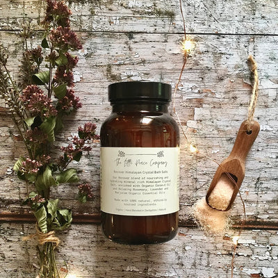 A jar of Recover Himalayan Crystal Bath Salts from The Little Peace Company, with a wooden scoop and dried herbs nearby, displayed on a wooden background.