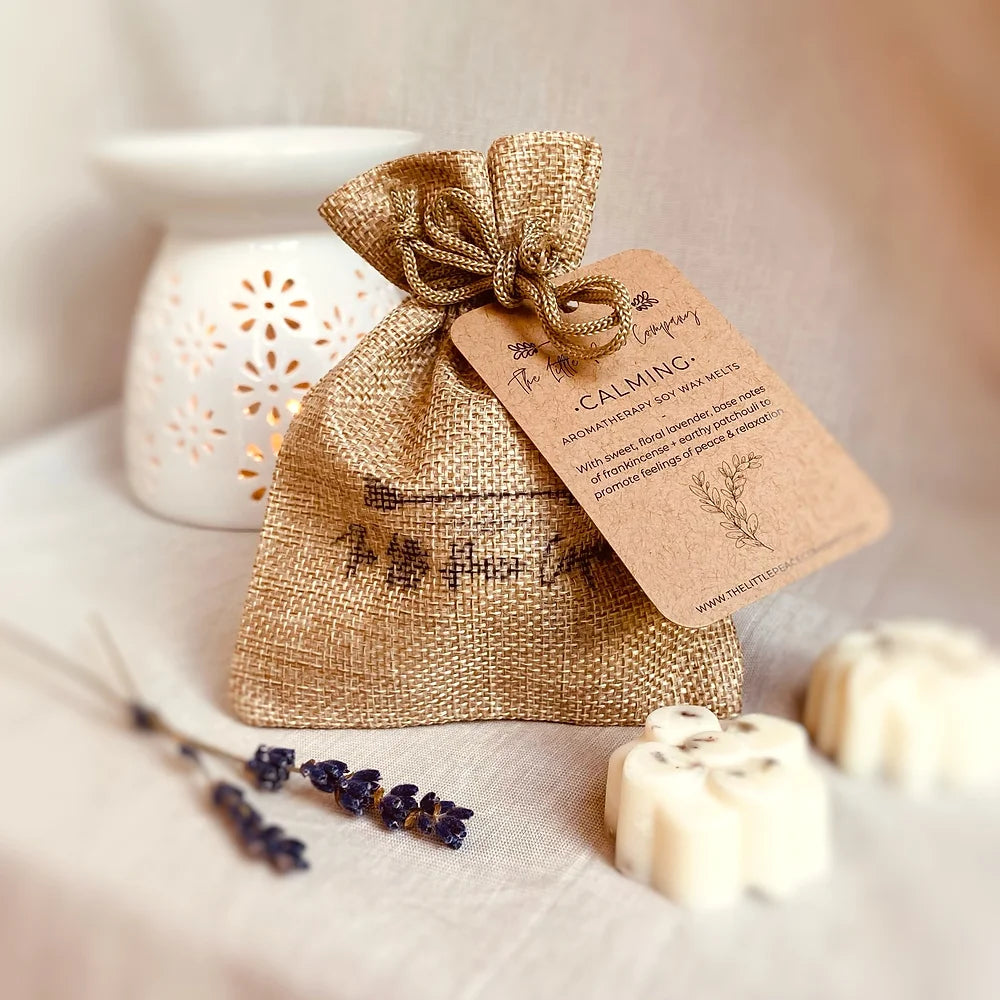 A burlap bag labeled 'Calming' containing large wax melts, alongside a white ceramic diffuser and dried lavender sprigs, all presented on a neutral background.