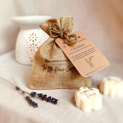 A burlap bag labeled 'Calming' containing large wax melts, alongside a white ceramic diffuser and dried lavender sprigs, all presented on a neutral background.