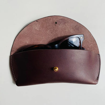 Brown leather sunglasses case with a gold button on a light gray background